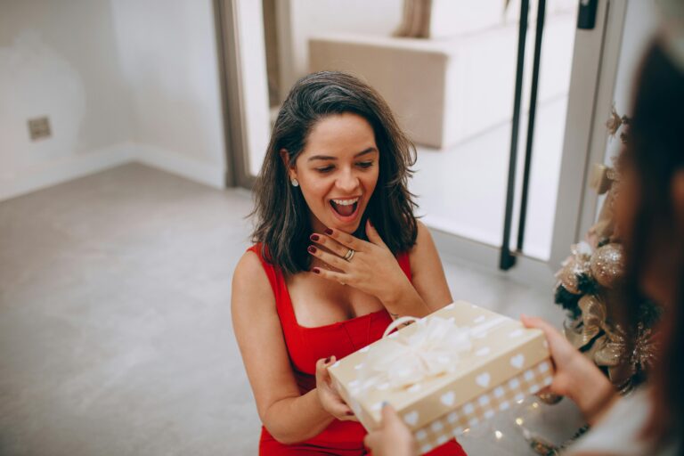 A woman in a red dress excitedly opens a beautifully wrapped Christmas gift indoors.